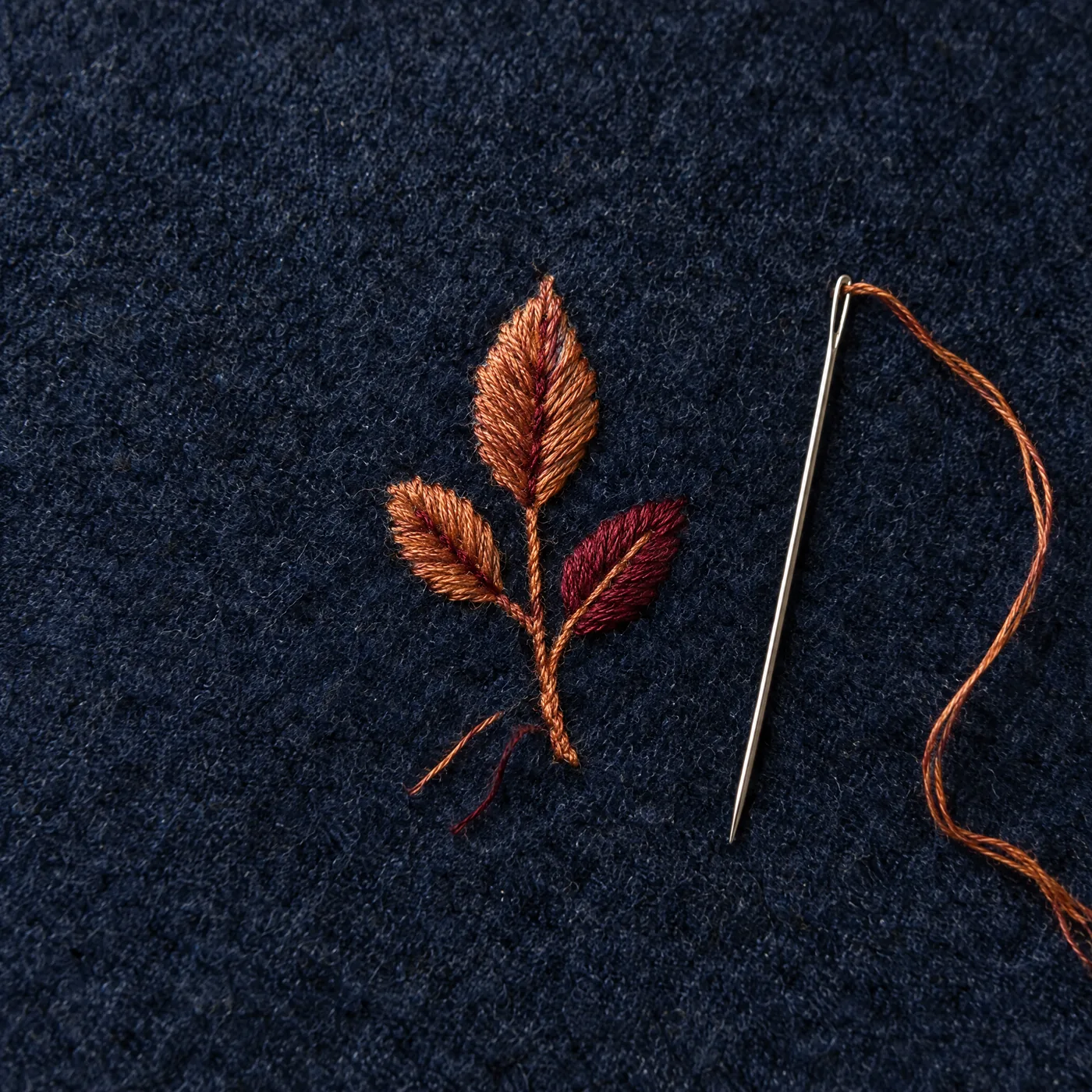 Brin de trois feuilles brodé à la main en fils cuivre et bordeaux sur une laine marine, à côté d'une aiguille enfilée